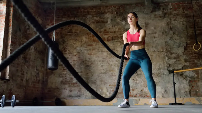 A woman wearing a pink sports bra and teal leggings exercises with battle ropes in an industrial-style gym, focused and engaged in an intense workout.