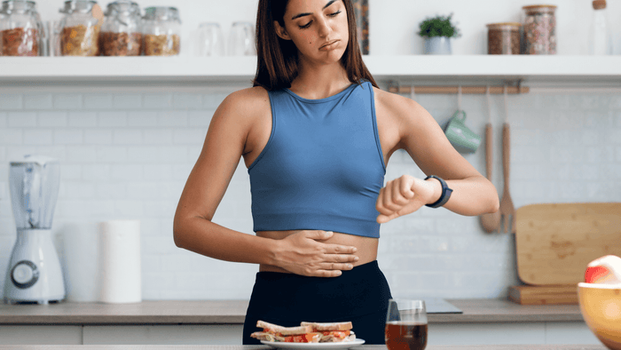 Woman in workout clothes holding her stomach and checking her watch in the kitchen.