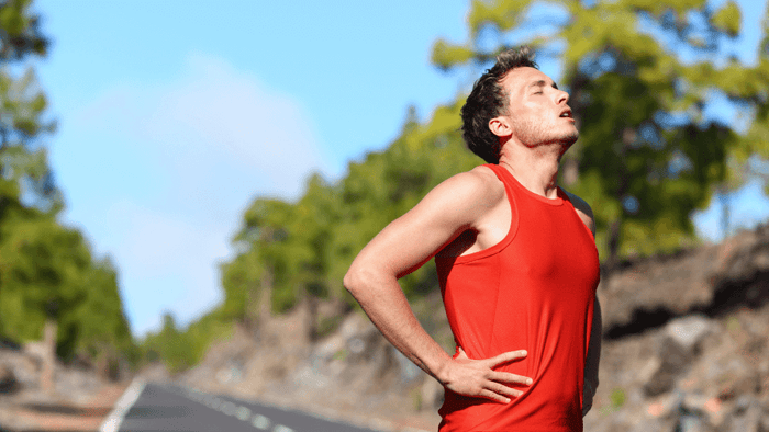 Male runner in a red tank top stands on a road with hands on hips, appearing fatigued or cramping during a workout.