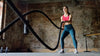 A woman wearing a pink sports bra and teal leggings exercises with battle ropes in an industrial-style gym, focused and engaged in an intense workout.
