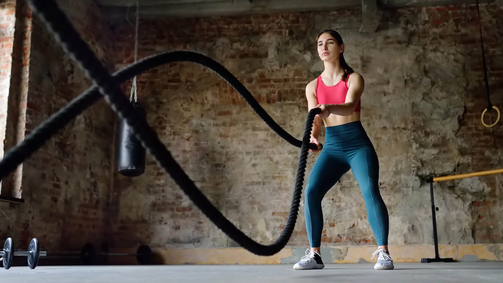 A woman wearing a pink sports bra and teal leggings exercises with battle ropes in an industrial-style gym, focused and engaged in an intense workout.