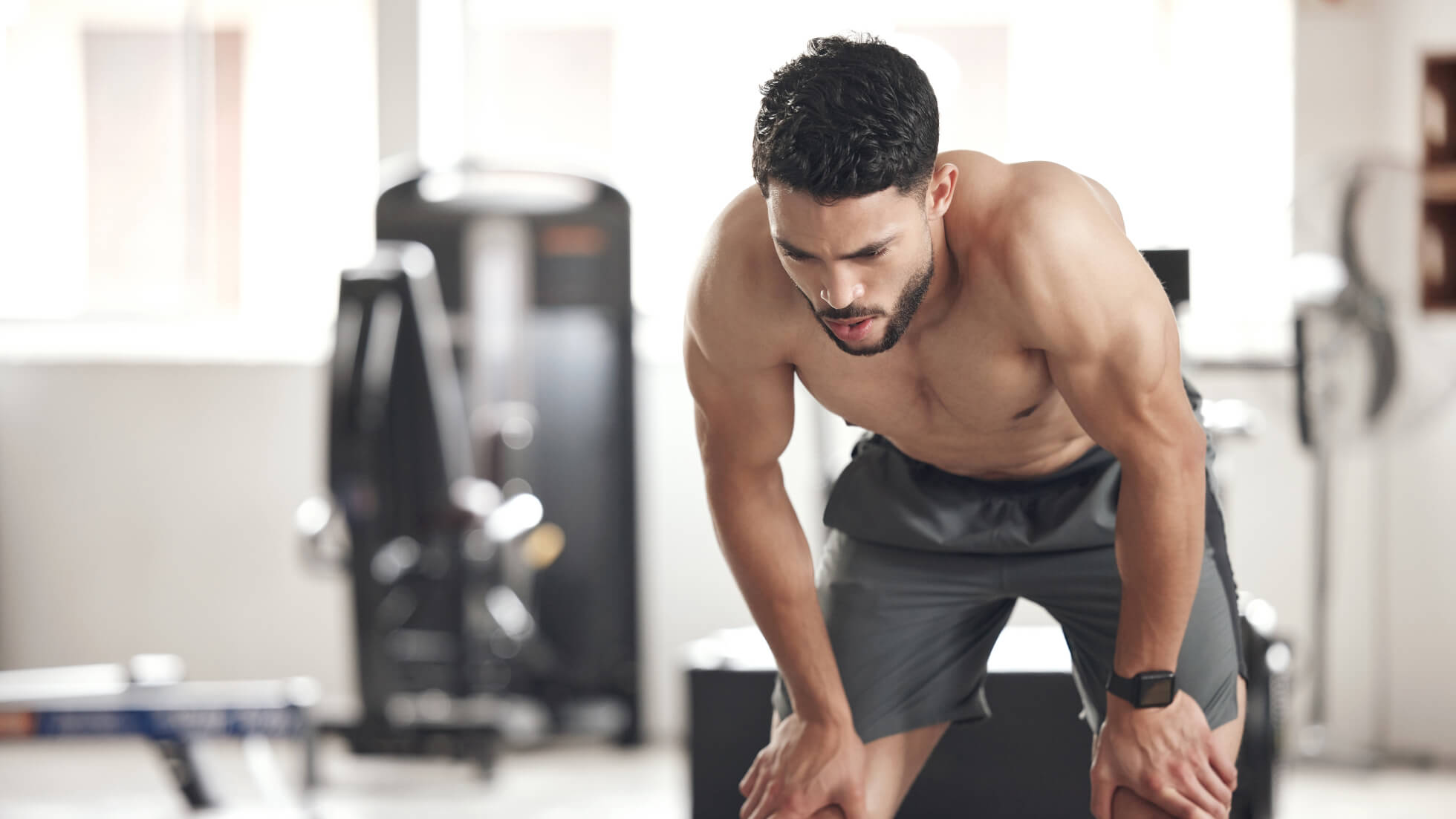 A shirtless man in workout shorts leans forward with hands on knees, appearing fatigued after exercise in a gym setting with equipment in the background.