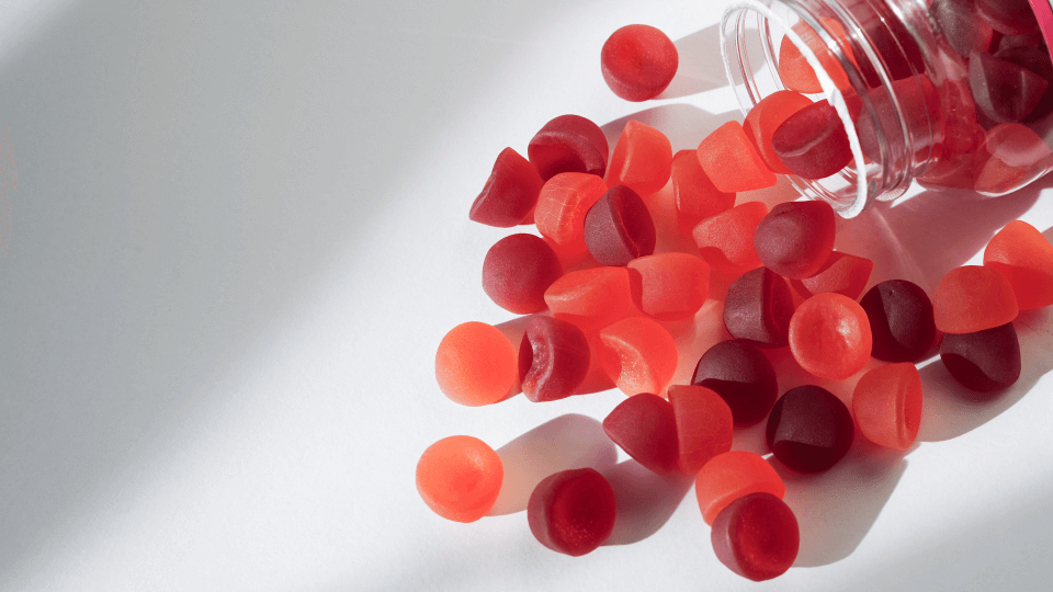 A clear jar tipped over with red and dark red gummy supplements spilling onto a white surface.