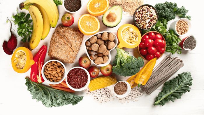 Assorted healthy foods including fruits, vegetables, grains, nuts, and legumes on a white table.
