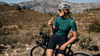Cyclist in helmet and sunglasses eating a snack while resting on a bike in a mountain landscape.