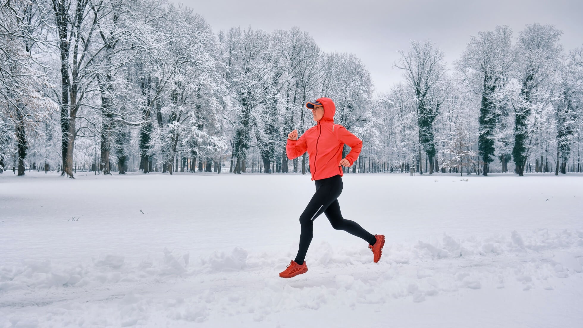 Runner in a red jacket jogs through a snowy park surrounded by snow-covered trees.