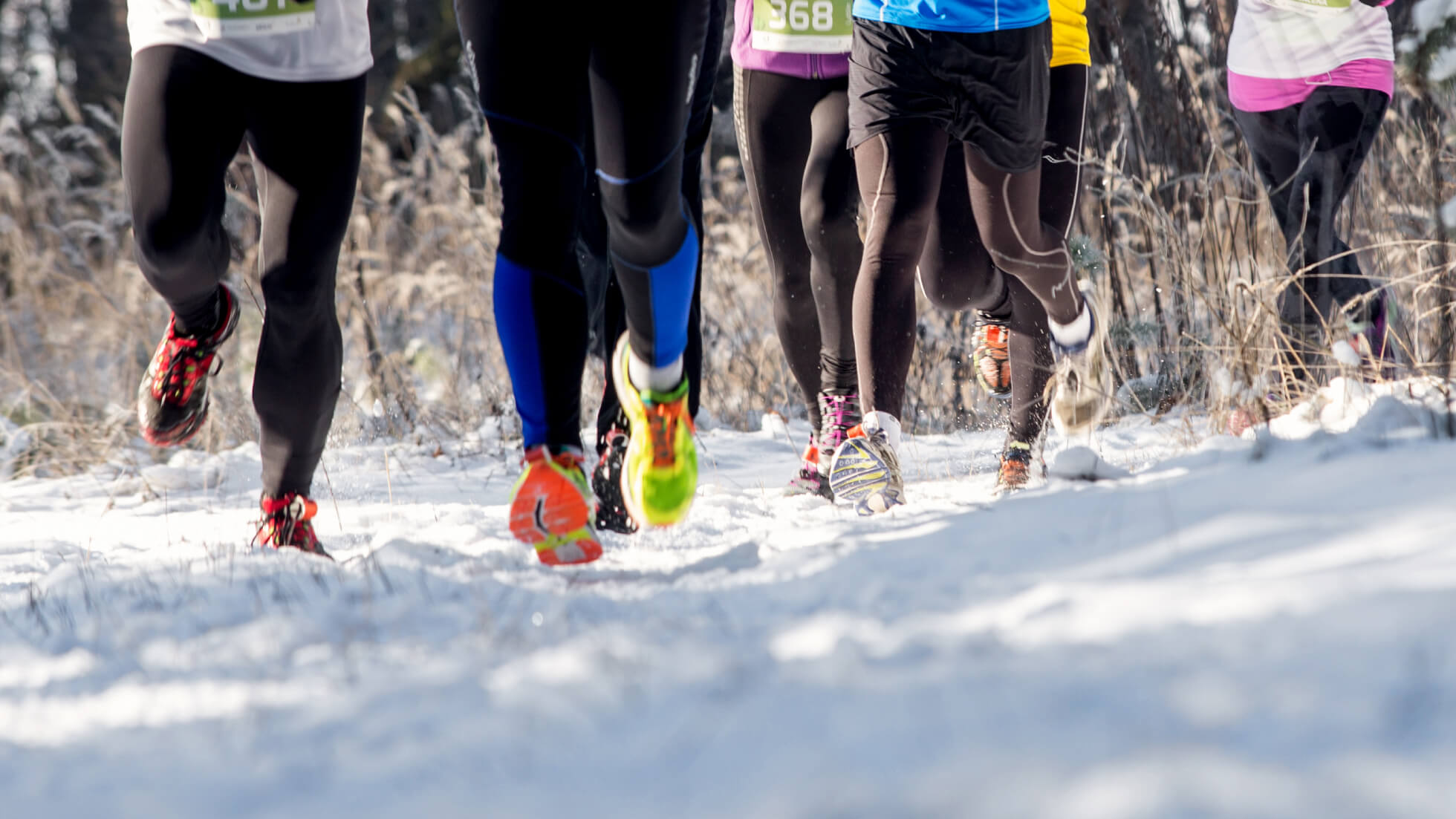 Runners wearing bright shoes and winter gear race along a snowy trail.