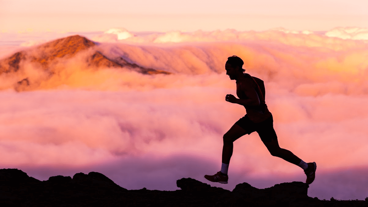 Runner silhouetted against a sunrise, moving along a rocky ridge above the clouds.