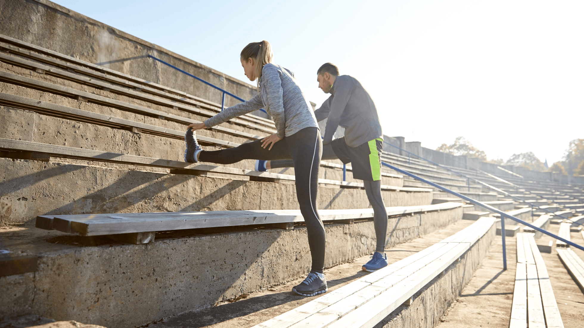 A man and a woman in athletic clothing stretch their legs on the steps of an outdoor stadium in the morning sunlight.