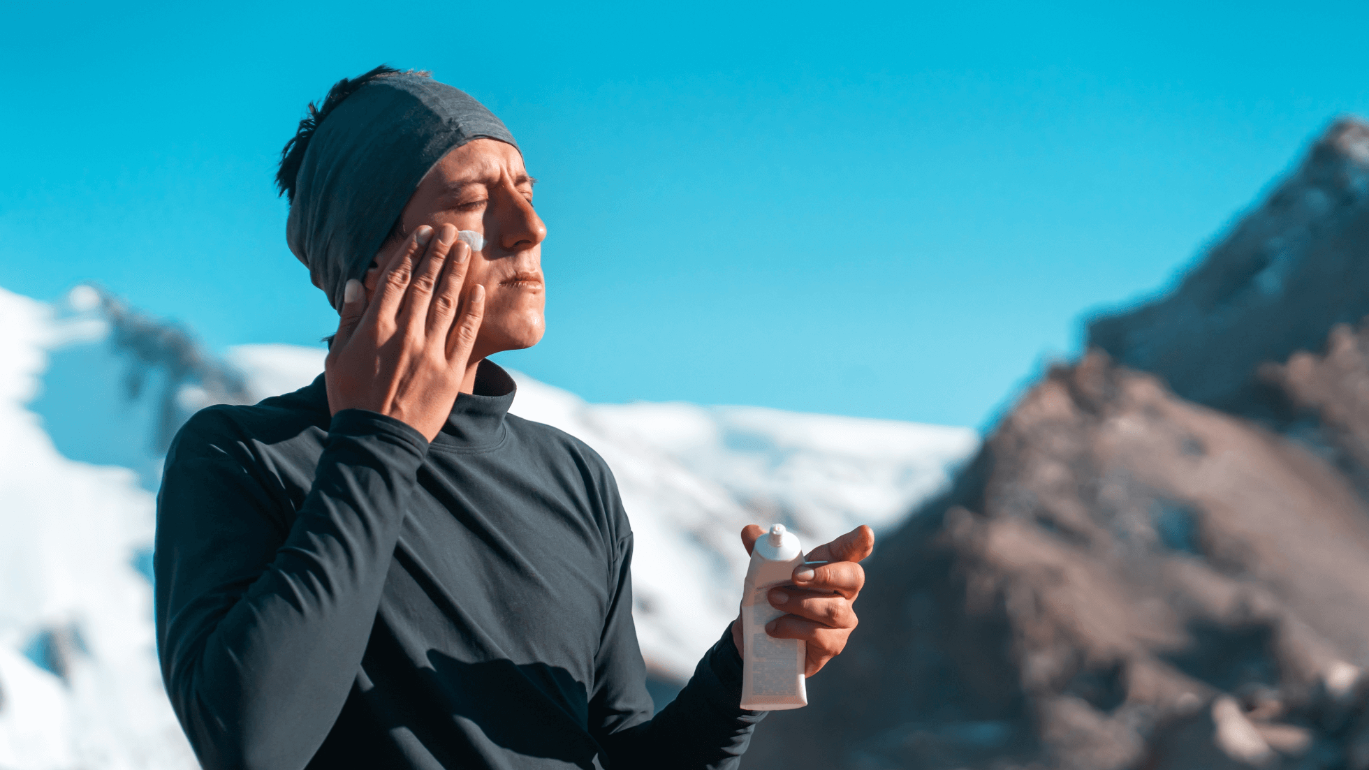 A person wearing a headband and long-sleeved shirt applies sunscreen to their face in a snowy mountain setting under a bright blue sky.