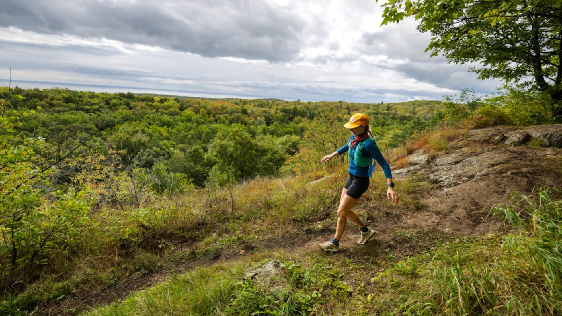 Trail runner wearing a yellow cap and blue vest running along a forested ridge.