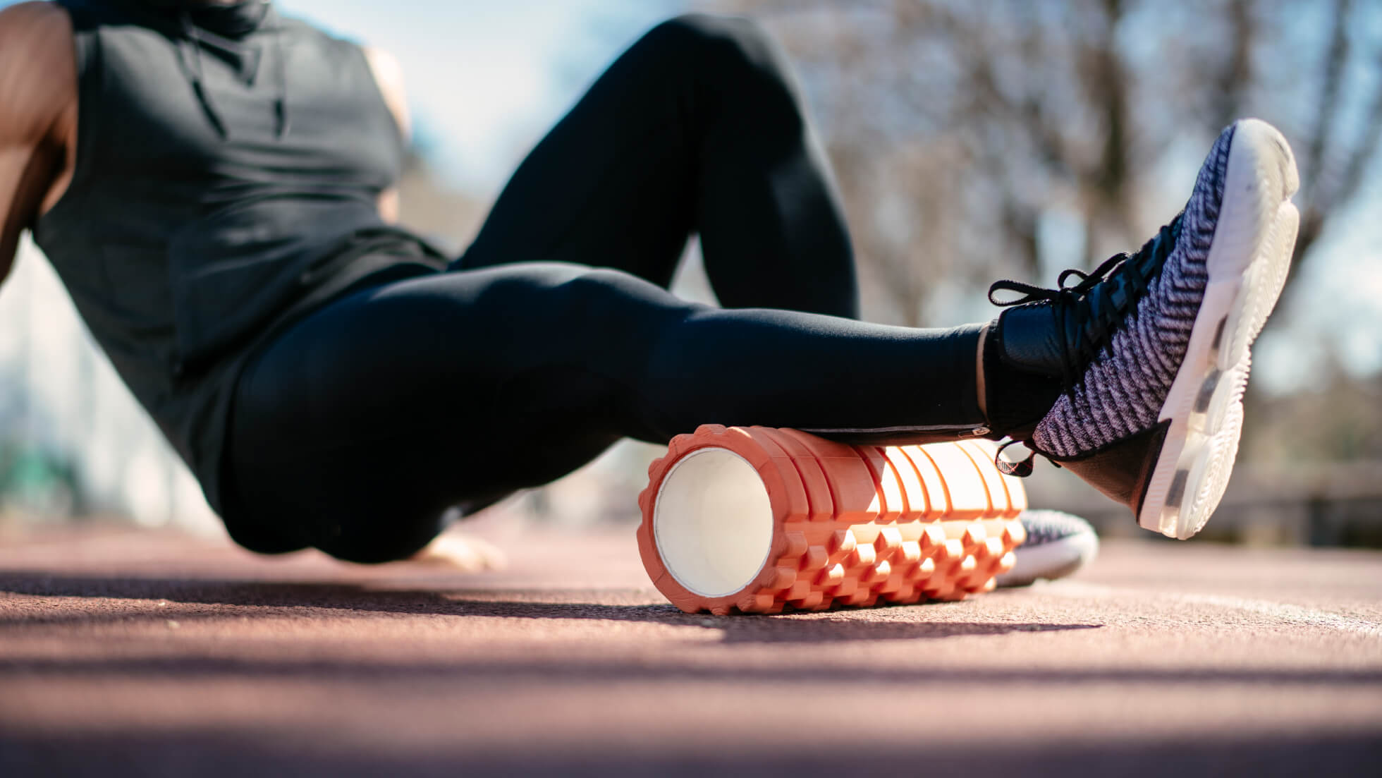 A person in athletic gear uses an orange foam roller on their thigh while outdoors, performing self-myofascial release as part of a recovery or warm-up routine.