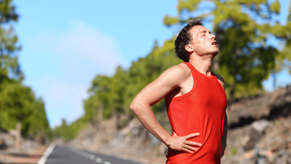 Male runner in a red tank top stands on a road with hands on hips, appearing fatigued or cramping during a workout.